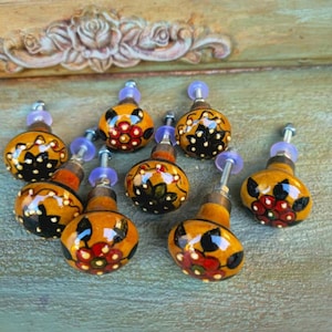 May include: A set of decorative cabinet knobs. Each knob has a yellow base with black, red, and white floral patterns. The knobs have metal stems and purple washers, ideal for adding a unique touch to furniture.