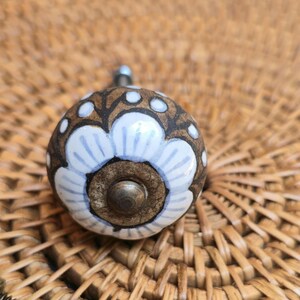 May include: A ceramic cabinet knob with a floral design. The knob has a brown base with white and pale blue floral accents. The knob is round and has a metal screw for attachment. The background is a woven wicker surface.