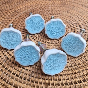 May include: Six ceramic cabinet knobs with a white and light blue cracked glaze design. The knobs have a silver metal screw and a clear plastic washer. The knobs are arranged on a woven wicker surface.