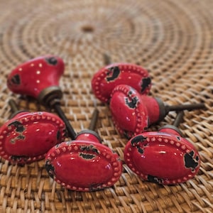 May include: A selection of red ceramic cabinet knobs with a distressed finish. The knobs have a beaded edge and black accents, with metal hardware. The knobs are oval and round shaped.