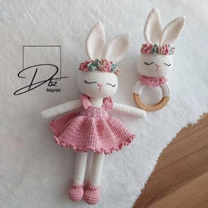 May include: Two handmade crochet bunny toys. One is a doll wearing a pink dress and shoes, the other is a rattle with a wooden ring. Both bunnies have white bodies, long ears, and floral headbands. The toys are on a white, textured surface.