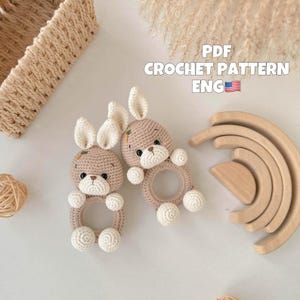 Puede incluir: Dos sonajeros de conejito de ganchillo en beige y blanco, con un anillo para agarrar y pompones blancos. La imagen también incluye un apilador de arcoíris de madera y el texto "PDF CROCHET PATTERN ENG".