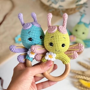 May include: Two handmade crochet insect baby rattles. One is light blue with purple accents, the other is yellow with pink accents. Both have wooden ring handles and small white flower details. The rattles are designed to be baby toys.