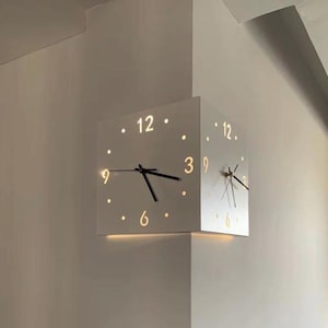 May include: A modern, white cube clock mounted on a corner wall. The clock has black hands and illuminated numbers at the 12, 3, 6, and 9 positions, with dots marking the other hours. The clock's design is minimalist and contemporary.