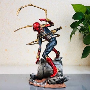 K&ouml;nnte beinhalten: Detaillierte Figur von Spider-Man in dynamischer Pose, mit rotem und goldenem Helm und Anzug mit blauen Akzenten. Die Figur ist auf einem strukturierten Metallsockel mit goldenen mechanischen Armen befestigt.