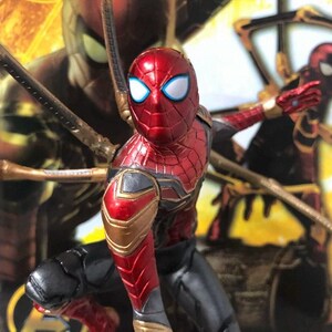 K&ouml;nnte beinhalten: Eine rot-goldene Spider-Man-Actionfigur mit wei&szlig;en Augen und schwarzen Akzenten. Die Figur befindet sich in einer dynamischen Pose, mit mechanischen Armen, die sich von ihrem R&uuml;cken aus erstrecken. Der Hintergrund zeigt ein verschwommenes Bild von Spider-Man.