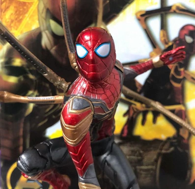 K&ouml;nnte beinhalten: Eine rot-goldene Spider-Man-Actionfigur mit wei&szlig;en Augen und schwarzen Akzenten. Die Figur befindet sich in einer dynamischen Pose, mit mechanischen Armen, die sich von ihrem R&uuml;cken aus erstrecken. Der Hintergrund zeigt ein verschwommenes Bild von Spider-Man.