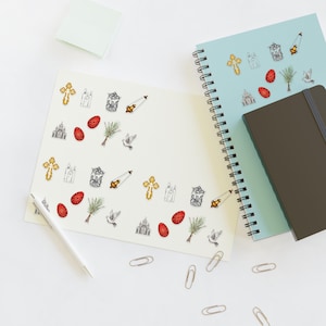 May include: A notebook and a journal with Easter-themed stickers. The stickers feature crosses, churches, decorated eggs, palm branches, doves, and candles. A pen and paperclips are also visible on the white surface.