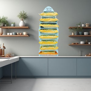 Könnte beinhalten: Ein hellblauer Aufsteller mit gelben und orangefarbenen Snackverpackungen. Der Ständer trägt oben den Text "Yum Yum Zeit". Der Ständer befindet sich in einer Küche mit Regalen und Schränken.