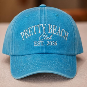 May include: A light blue baseball cap with the text "PRETTY BEACH Club EST. 2026" embroidered in white. The cap has a curved brim and a fabric-covered button on top. The hat is made of a soft, textured material.