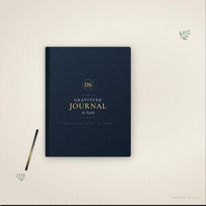 Puede incluir: Un diario de gratitud azul marino con letras doradas y un bolígrafo dorado. La portada del diario dice "Gratitude Journal & Faith" con las iniciales "DS" en un círculo. El diario es de Davanic Studio.