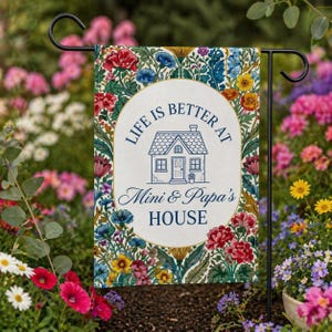 May include: A garden flag with the text "LIFE IS BETTER AT Mini & Papa's HOUSE" in a white oval, surrounded by a floral design with red, blue, and yellow flowers. The flag is displayed in a garden setting.