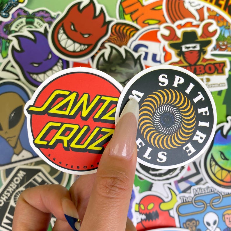 May include: A collection of colorful skateboard stickers. Two round stickers are in focus: one with "SANTA CRUZ" in yellow letters on a red background, and another with "SPITFIRE" around a spiral design. Other stickers feature various graphics.