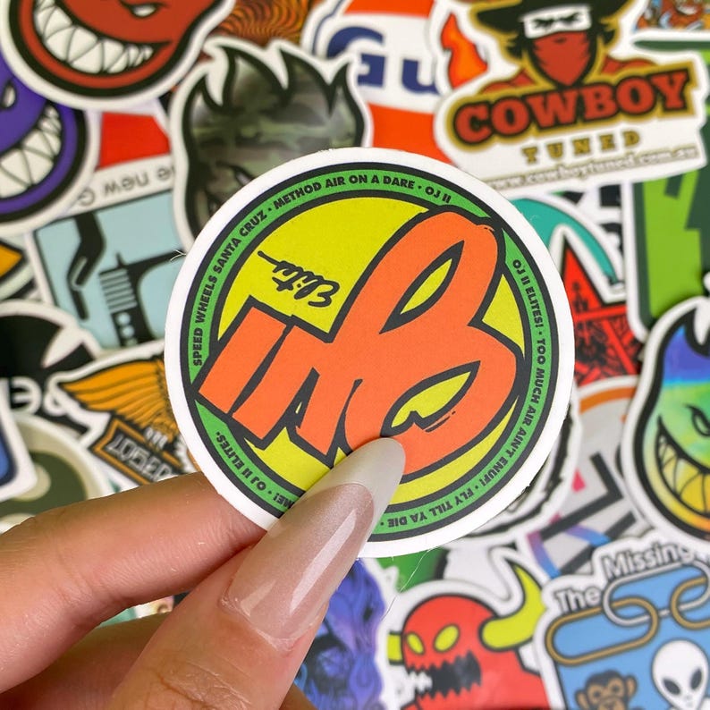 May include: A round sticker with the letters "MB" in orange, outlined in black, on a yellow and green background. The sticker has the text "SPEED WHEELS SANTA CRUZ" and "OJI ELITES" around the edges. Other stickers are in the background.