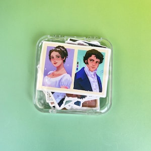 May include: A clear plastic case containing stickers with illustrations of two people. One sticker features a woman in a white dress and the other a man in a blue coat. The stickers are on a green background.