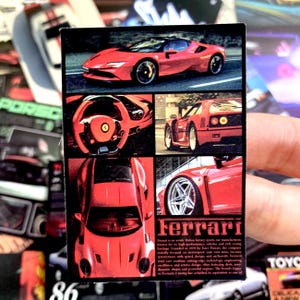 May include: A rectangular sticker featuring a collage of red Ferrari sports cars. The design includes a side view, a steering wheel interior, a rear view, a top-down view, and a close-up of a wheel. The text Ferrari is printed at the bottom.
