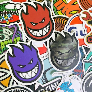 May include: A collection of colorful stickers featuring various designs. The prominent stickers showcase a grinning, flame-shaped face in red, purple, and camouflage patterns. Other stickers include skate-related graphics and logos.