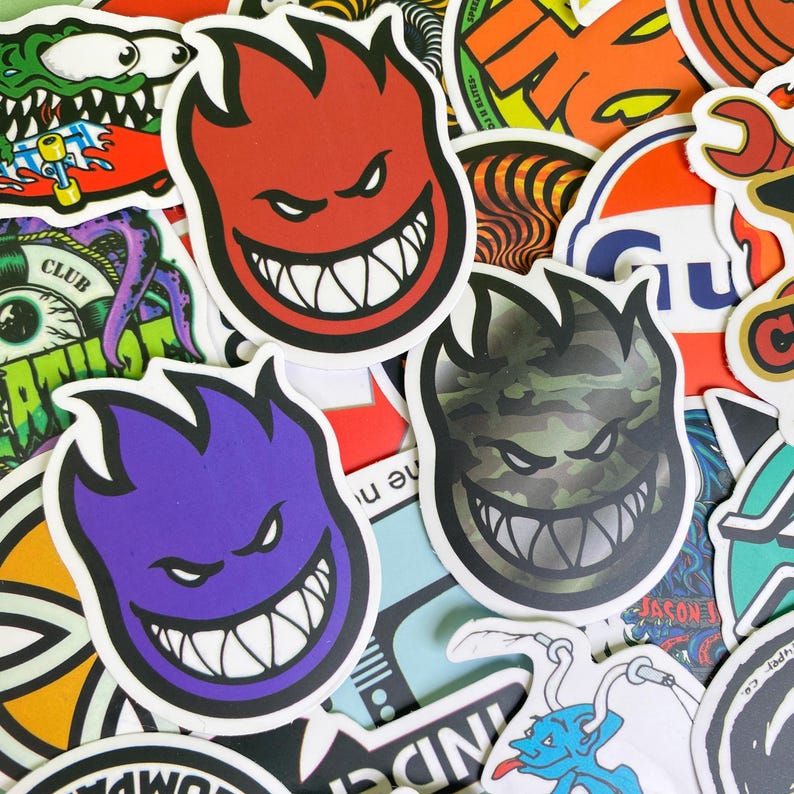 May include: A collection of colorful stickers featuring various designs. The prominent stickers showcase a grinning, flame-shaped face in red, purple, and camouflage patterns. Other stickers include skate-related graphics and logos.