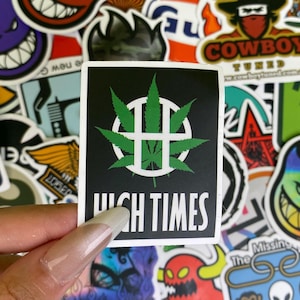 May include: A square sticker with a black background features a green cannabis leaf design inside a white circle with a cross. The words "HIGH TIMES" are printed in white below the design. The sticker is surrounded by other colorful stickers.