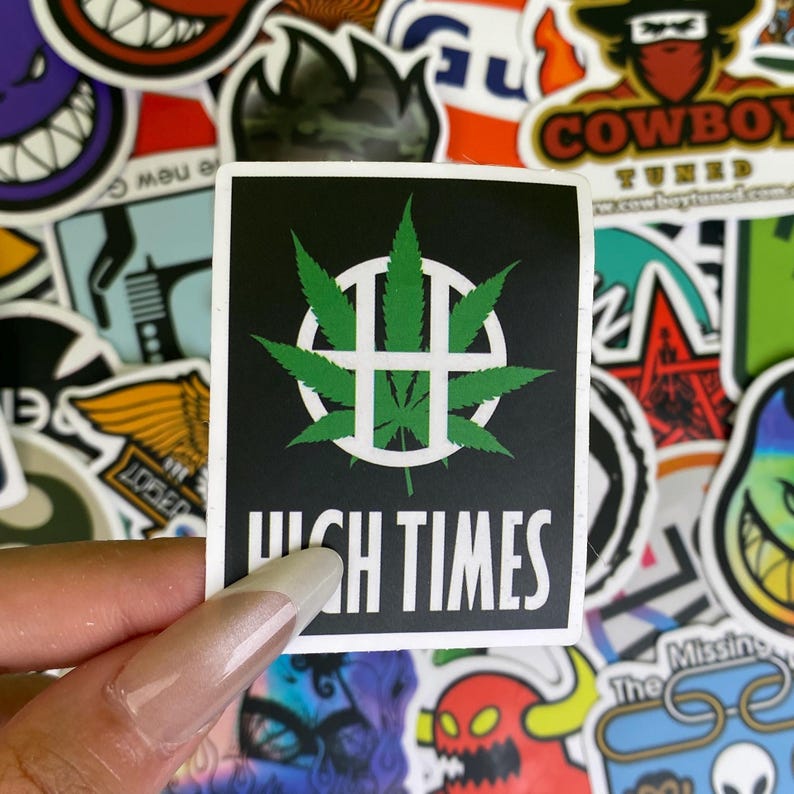 May include: A square sticker with a black background features a green cannabis leaf design inside a white circle with a cross. The words "HIGH TIMES" are printed in white below the design. The sticker is surrounded by other colorful stickers.