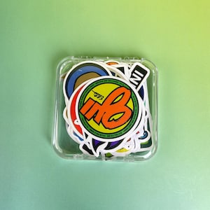 May include: A clear plastic case filled with colorful stickers. The stickers feature various designs, including a large orange and yellow "MB" logo, and text that reads "SUPER WHEEL SANTA CRUZ". The background is a solid light green.