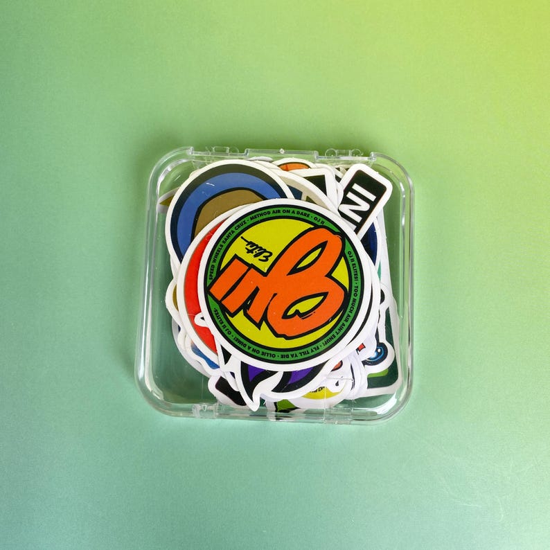 May include: A clear plastic case filled with colorful stickers. The stickers feature various designs, including a large orange and yellow "MB" logo, and text that reads "SUPER WHEEL SANTA CRUZ". The background is a solid light green.
