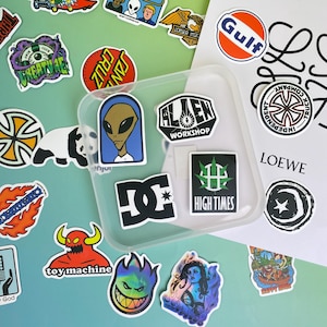May include: A collection of colorful stickers with various designs, including an alien, a panda, and logos. Some stickers feature text like "Alien Workshop" and "High Times". The stickers are arranged on a light green and blue surface.