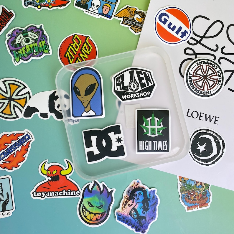 May include: A collection of colorful stickers with various designs, including an alien, a panda, and logos. Some stickers feature text like "Alien Workshop" and "High Times". The stickers are arranged on a light green and blue surface.