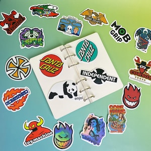 May include: A collection of colorful stickers featuring various designs, including logos, cartoon characters, and text. Stickers include the Santa Cruz logo, a panda, and the text "Gone Fishing". The stickers are arranged on a white notebook.