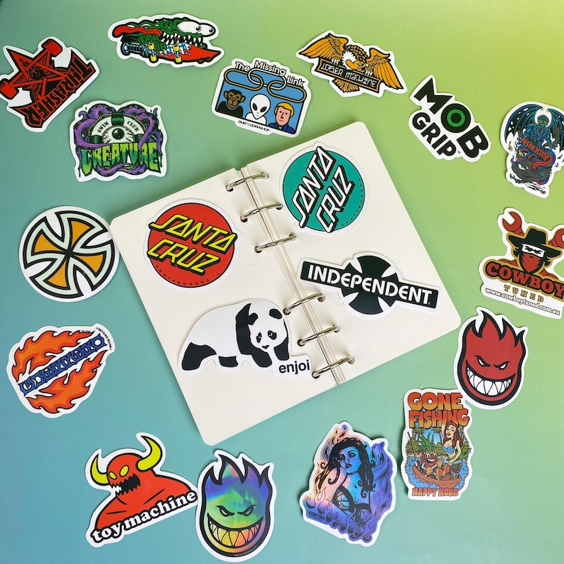 May include: A collection of colorful stickers featuring various designs, including logos, cartoon characters, and text. Stickers include the Santa Cruz logo, a panda, and the text "Gone Fishing". The stickers are arranged on a white notebook.