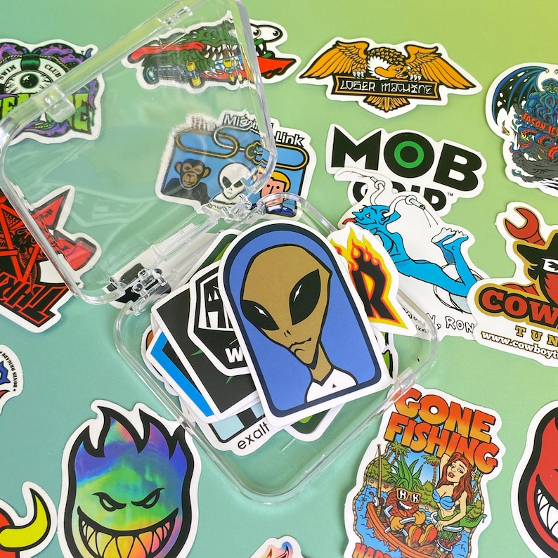 May include: A clear phone case filled with colorful stickers. The stickers feature various designs, including an alien with a blue head covering, a cartoon eagle, and the words "MOB" and "GONE FISHING".
