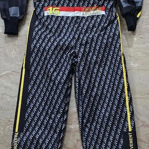 Puede incluir: Un mono de carreras negro y gris con detalles amarillos, con logotipos de patrocinadores y el logotipo de HP. El mono incluye el número 16 y el nombre CHARLES LECLERC. Las palabras "PASSION AND ACHIEVEMENT" están impresas en las piernas.