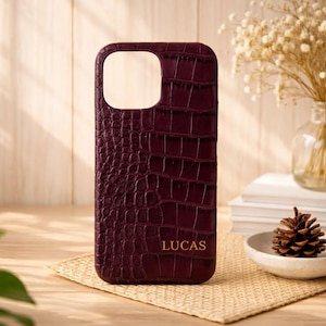 May include: A burgundy phone case with a crocodile skin texture. The case has a cutout for the camera and the name "LUCAS" in gold lettering. The case is on a woven mat with a wooden background.