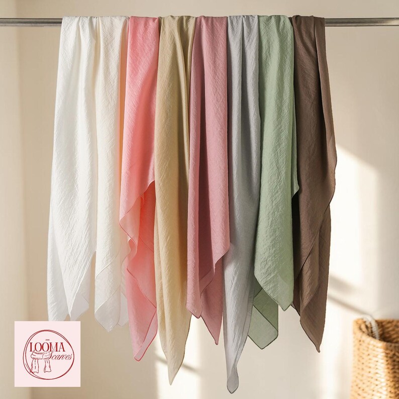 May include: A collection of seven rectangular scarves hanging on a metal rod, featuring a variety of solid colours including white, light pink, beige, dusty rose, light grey, sage green, and brown. Each scarf has a textured, lightweight fabric finish.