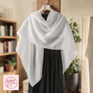 May include: A lightweight white scarf with a subtle crinkled texture draped over a black sleeveless dress. The scarf is displayed on a hanger and features a logo in the corner with the text LOOMA Scarves and an illustration of a scarf.