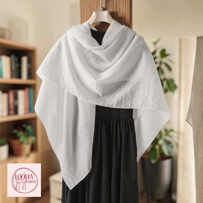 May include: A lightweight white scarf with a subtle crinkled texture draped over a black sleeveless dress. The scarf is displayed on a hanger and features a logo in the corner with the text LOOMA Scarves and an illustration of a scarf.