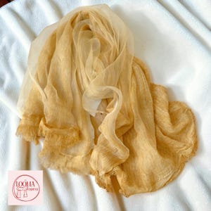 May include: A sheer, golden-yellow scarf with a lightweight, crinkled texture and frayed edges. The fabric is translucent and draped in soft folds. A small square logo in the corner features the text LOOMA Scarves with a line drawing of a scarf.