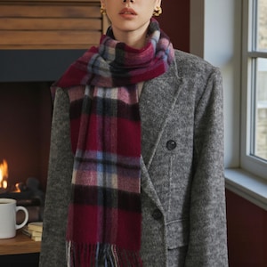 May include: A tartan scarf in burgundy, black, and blue is worn over a grey coat. The scarf is fringed and wrapped around the neck. The coat is a double-breasted style, offering a classic look. The scarf's pattern is a traditional plaid.