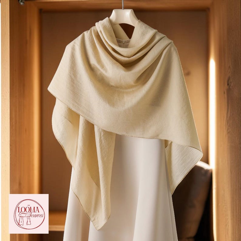 May include: A light cream-coloured scarf draped over a white garment on a hanger. The scarf features a soft, textured fabric with visible folds. A logo in the bottom left corner displays the text LOOMA Scarves with a line drawing of a scarf.