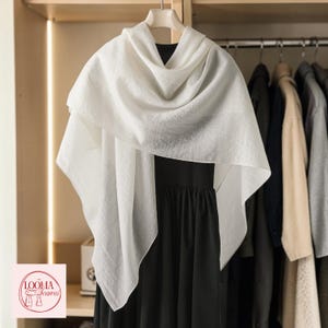 May include: A solid white, lightweight, semi-sheer rectangular scarf draped over a black garment. The scarf features a subtle, fine-textured weave throughout its surface. A small logo in the corner displays the text LOOMA Scarves with an illustration.