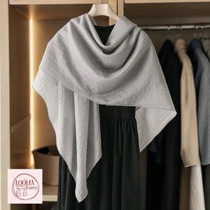 May include: A light grey, textured scarf draped over a black garment on a hanger. The scarf features a soft, crinkled fabric appearance with clean edges. A small square logo in the corner displays the text LOOMA Scarves with a line drawing.