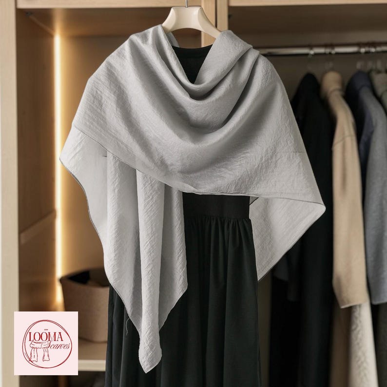 May include: A light grey, textured scarf draped over a black garment on a hanger. The scarf features a soft, crinkled fabric appearance with clean edges. A small square logo in the corner displays the text LOOMA Scarves with a line drawing.