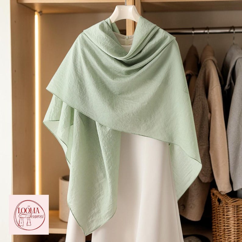 May include: A light sage green scarf draped over a white garment on a hanger. The scarf features a soft, textured fabric with visible folds and a subtle sheen. A small square logo in the corner displays the text LOOMA Scarves with a line drawing.