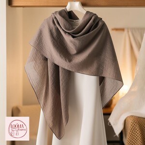 May include: A taupe-coloured scarf with a textured, lightweight fabric draped over a white garment. The scarf features a soft, matte finish and hangs in loose, flowing folds. A circular logo with the text LOOMA Scarves is visible in the corner.