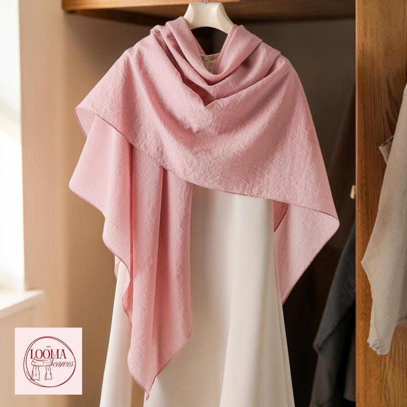May include: A soft, light pink scarf draped over a white garment on a hanger. The scarf features a textured, slightly wrinkled fabric finish and is displayed in a way that shows its folds and drape. A logo with the text LOOMA Scarves is visible.
