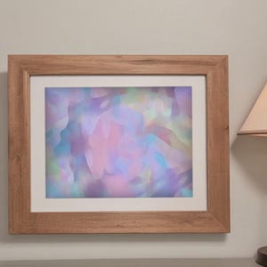 Puede incluir: Una impresión artística abstracta enmarcada con efecto acuarela. La obra presenta una mezcla de colores pastel, incluyendo rosa, morado y azul, creando una estética suave y soñadora. El marco es de madera clara.