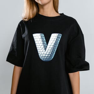May include: Black t-shirt featuring a large, 3D letter "V" with a golf ball texture. The white and gray design creates a striking contrast against the dark fabric. The shirt has short sleeves and a relaxed fit.