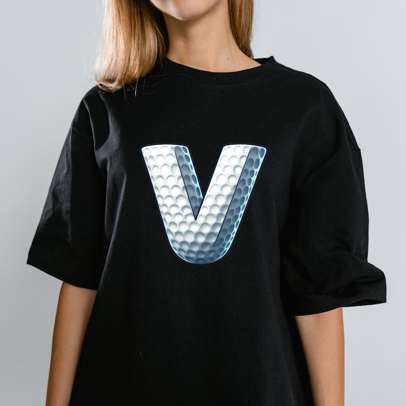 May include: Black t-shirt featuring a large, 3D letter "V" with a golf ball texture. The white and gray design creates a striking contrast against the dark fabric. The shirt has short sleeves and a relaxed fit.