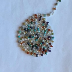 May include: A long necklace featuring a variety of colorful gemstones, including turquoise, amethyst, and orange stones, strung on a delicate silver chain. The necklace is displayed on a white surface.