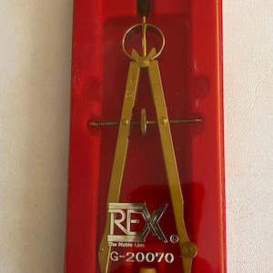 May include: A gold-colored compass set in a red plastic case. The compass is labeled "REX" and "G-20070". The case also includes text indicating the product is from Rex Graphic Supply, a division of American Pin & Fastener Corp.